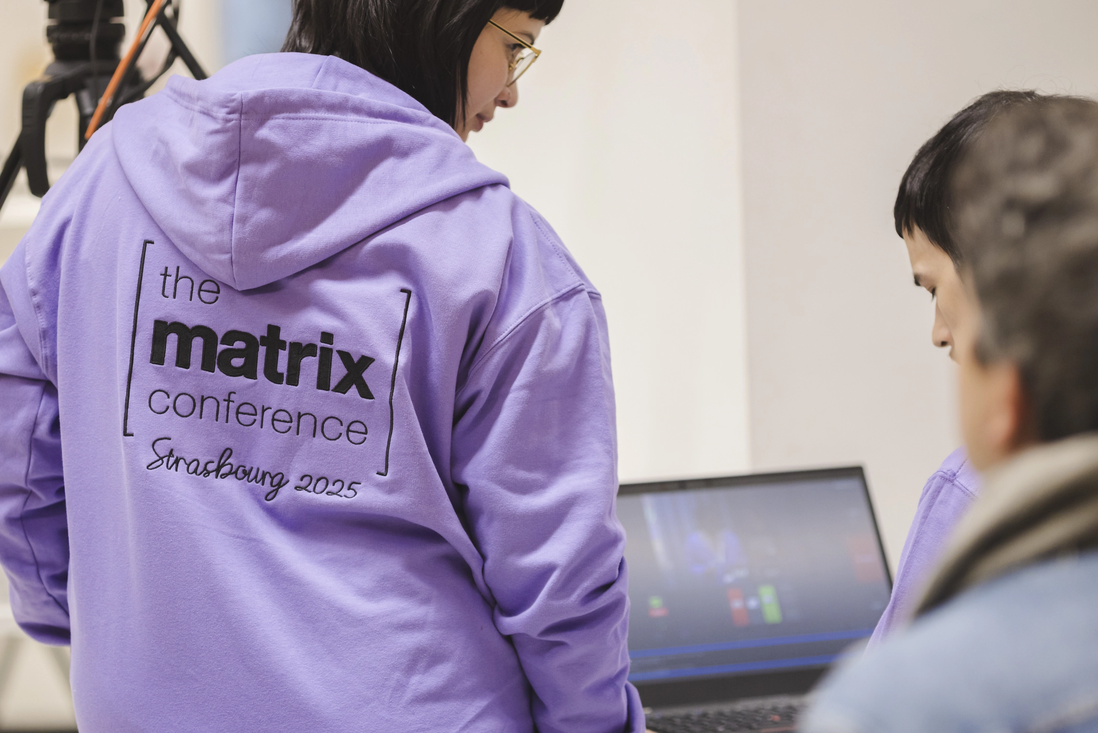 A photo of a woman wearing a purple hoodie with the logo of The Matrix Conference embroidered prominently. She is looking at another volunteer, smiling. The other volunteer is operating video mixing software on a laptop.